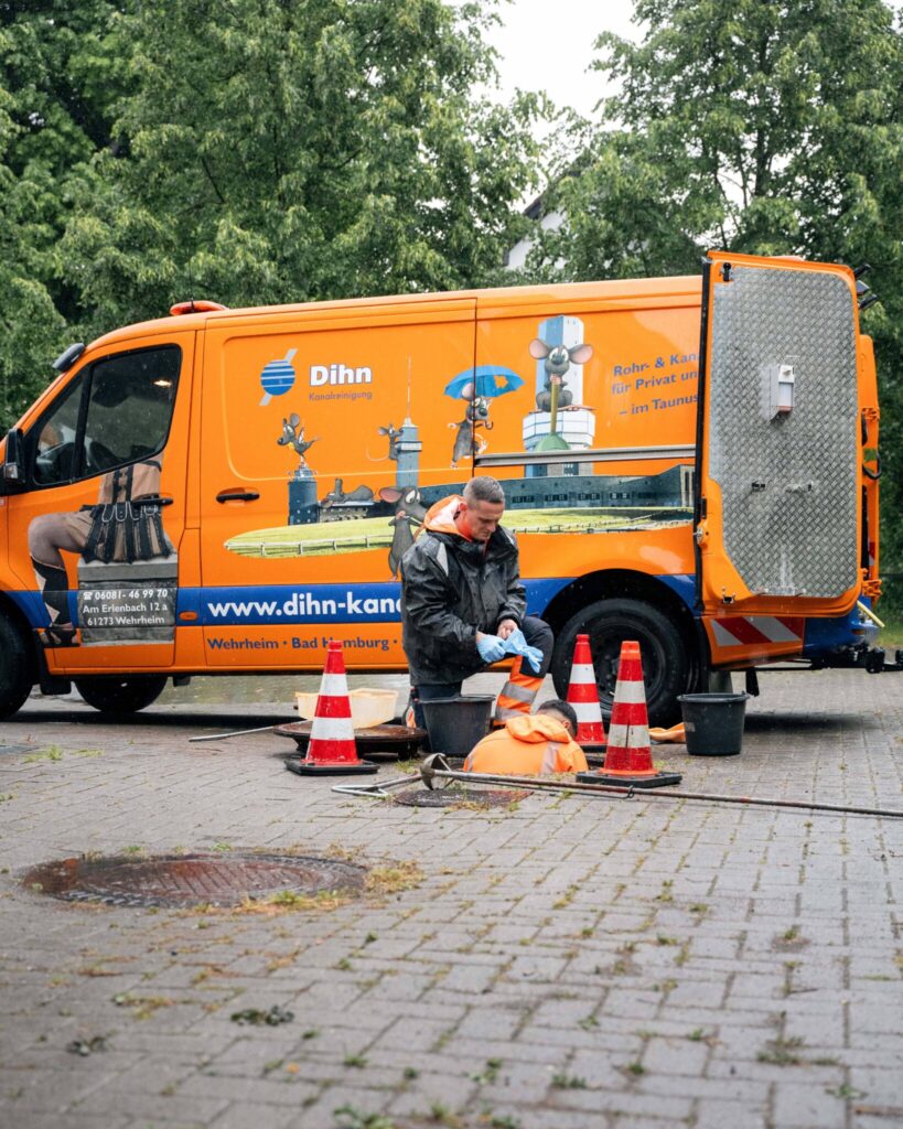 Ein Mitarbeiter von Dihn Kanalreinigung bei der Arbeit vor dem orangefarbenen Einsatzfahrzeug. Neben ihm zwei Eimer und drei Pylonen.