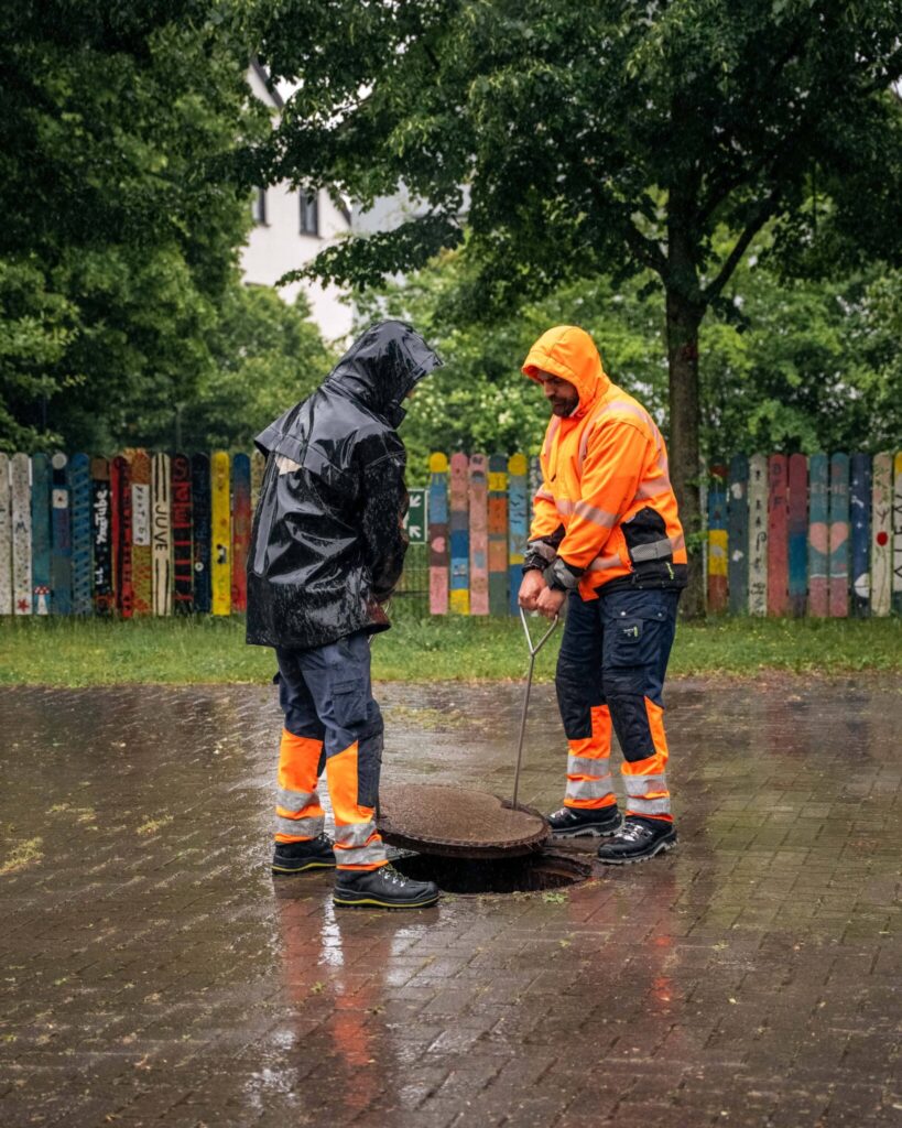 Zwei Mitarbeiter von Dihn Kanalreinigung in Orange und Schwarz, die bei Regen einen Kanaldeckel anheben.